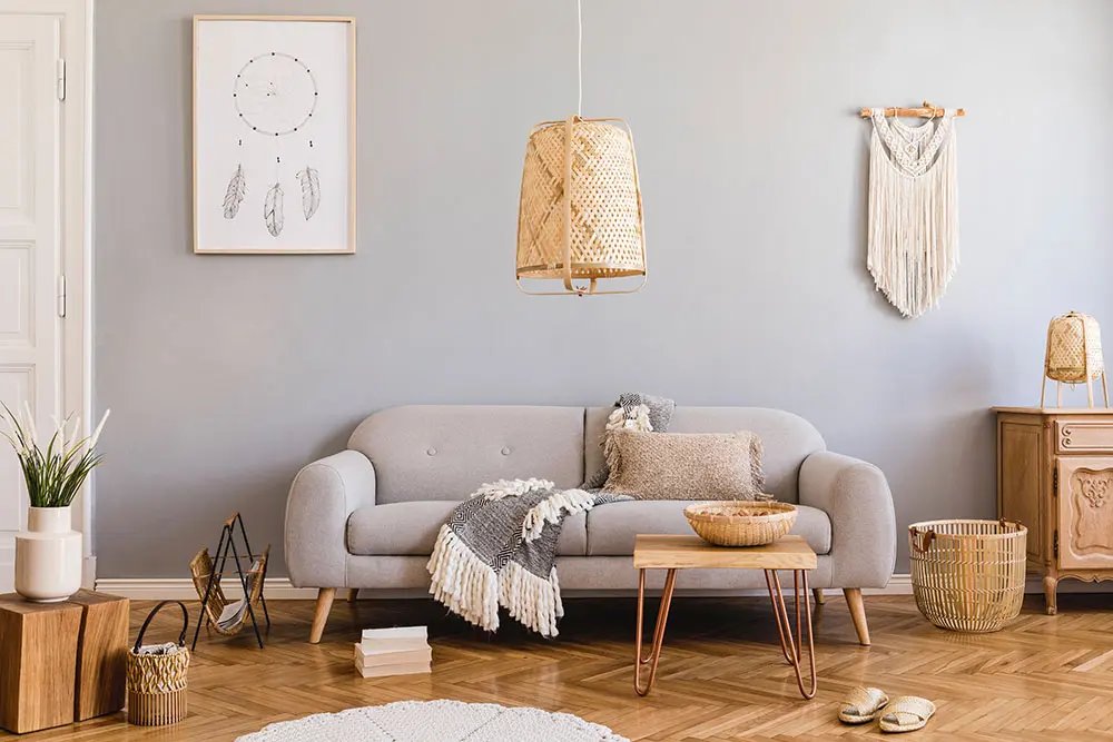 Stylish and design home interior of living room with gray sofa, wooden cube, commode, pillow, macrame, rattan lamp, basket, plants and elegant accessories.